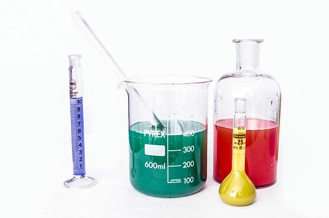 Photo of a small graduated cylinder, large beaker with glass stirring rod, large bottle, and small volumetric flask containing different colored liquids.
